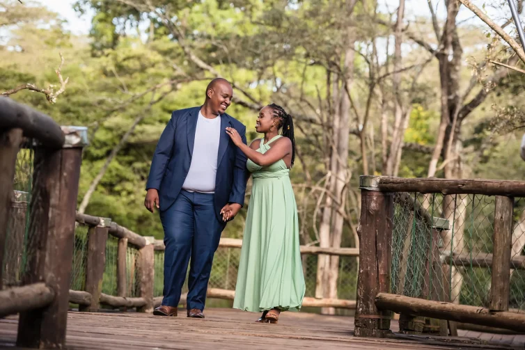 A couple walking hand-in-hand on a wooden bridge surrounded by green trees.